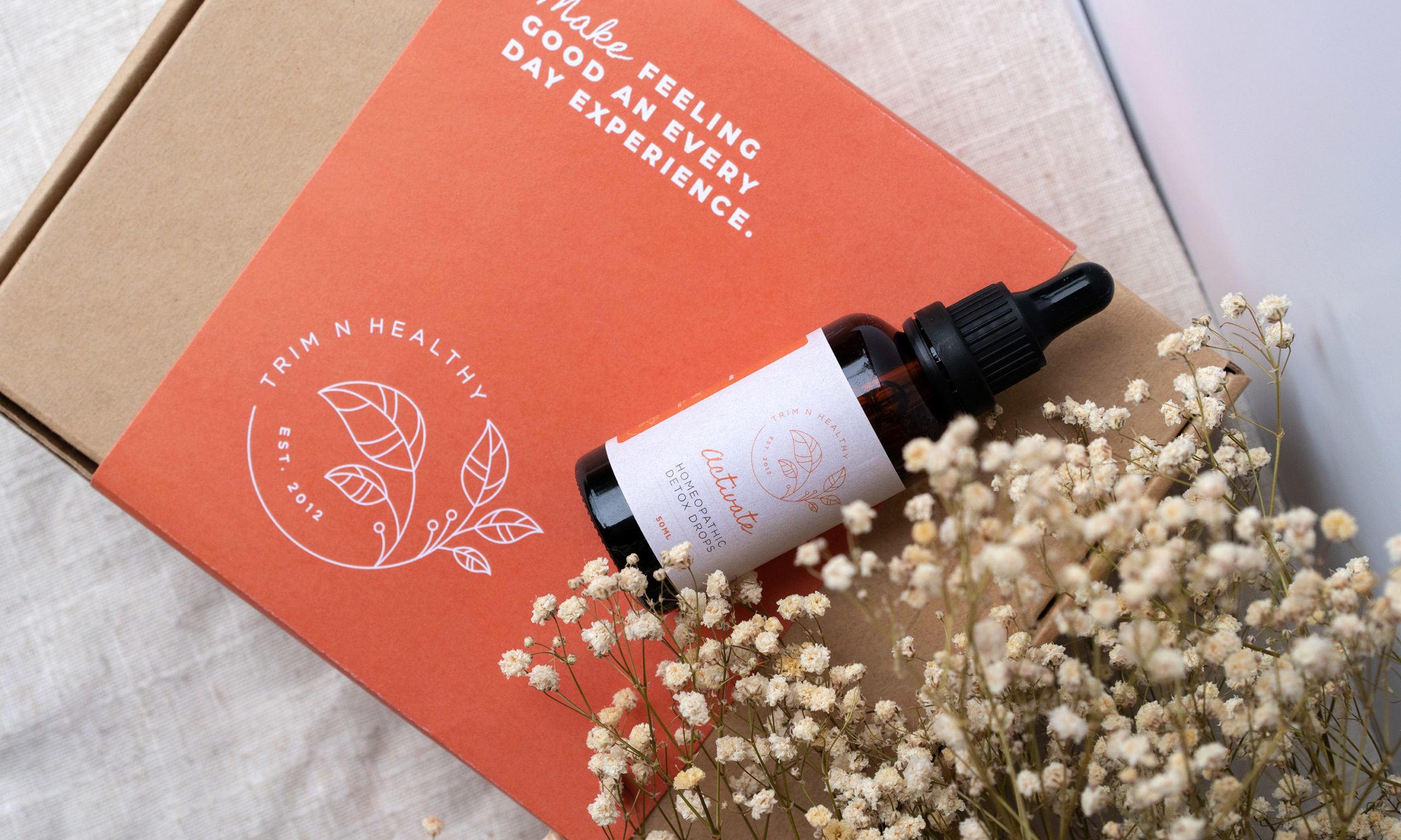 Trim N Healthy natural weight loss Activate drops with white flowers and a coral coloured card that reads Make feeling good an every day experience.